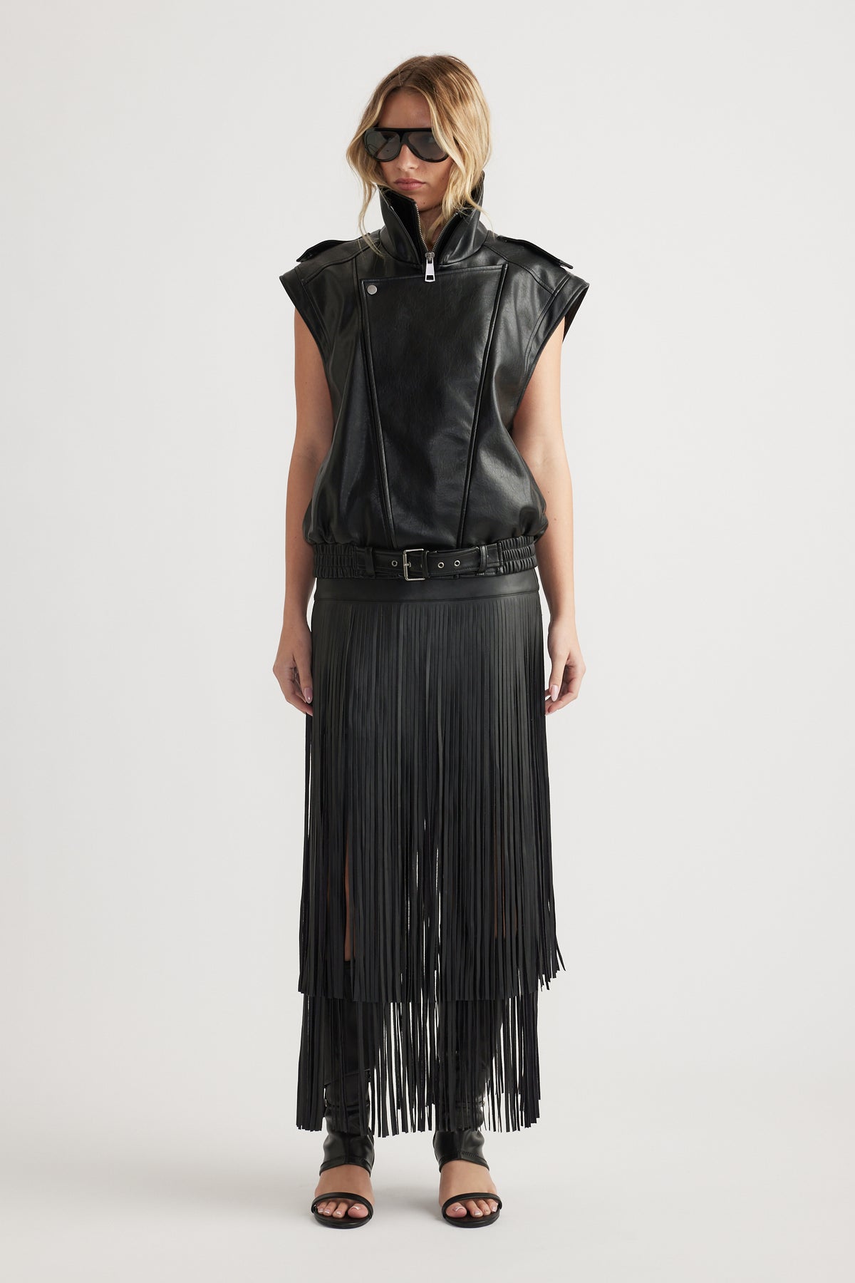 Woman wearing a belted black faux leather vest paired with a long fringed skirt and open-toe heels