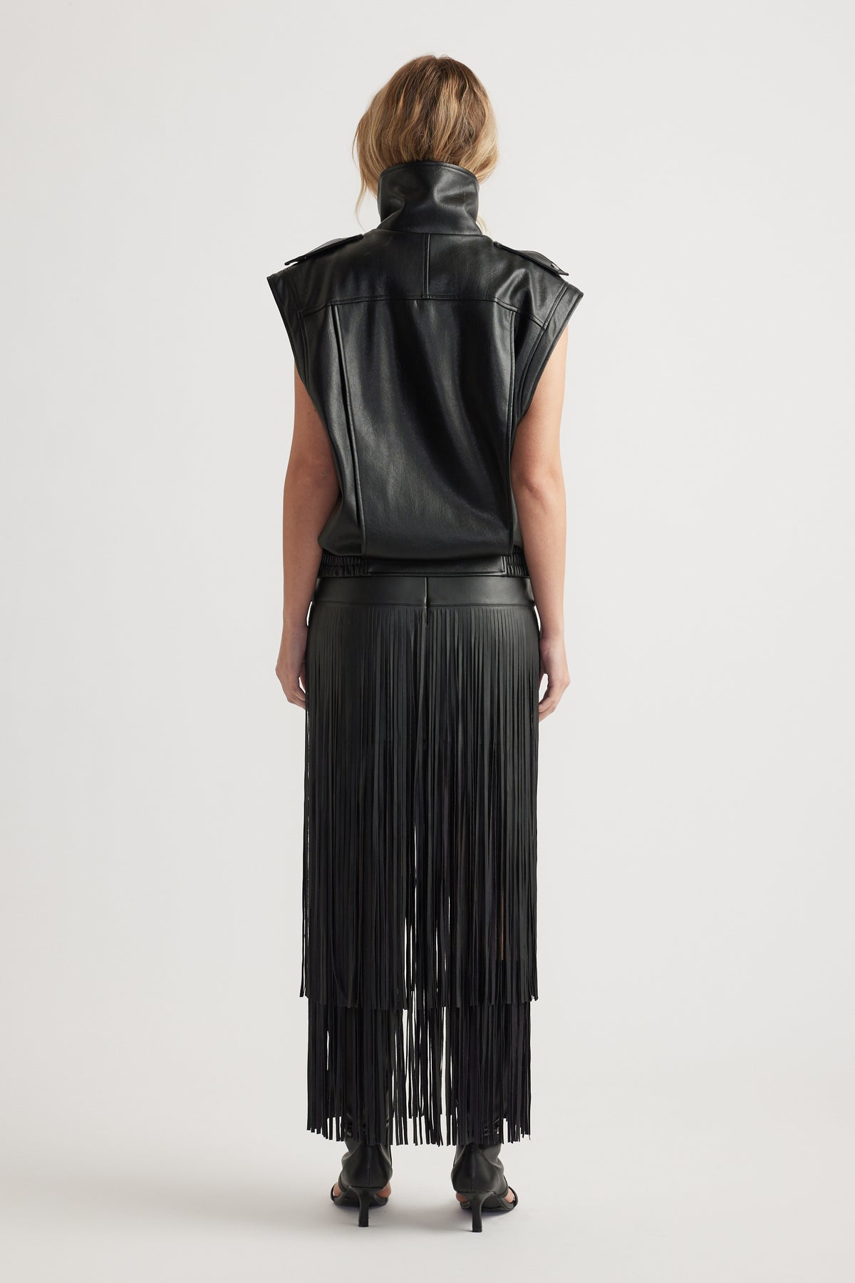 Back view of woman wearing a black faux leather vest paired with a long fringed skirt and heeled boots