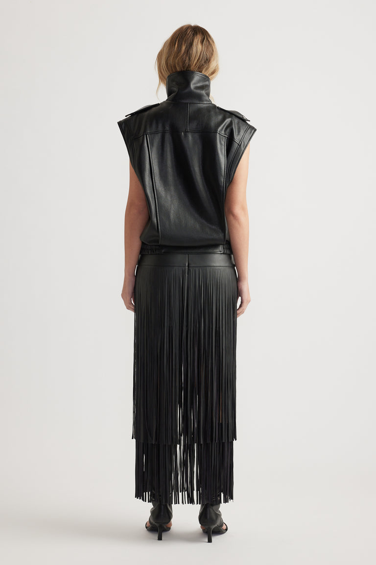 Back view of woman wearing a black faux leather vest paired with a long fringed skirt and heeled boots
