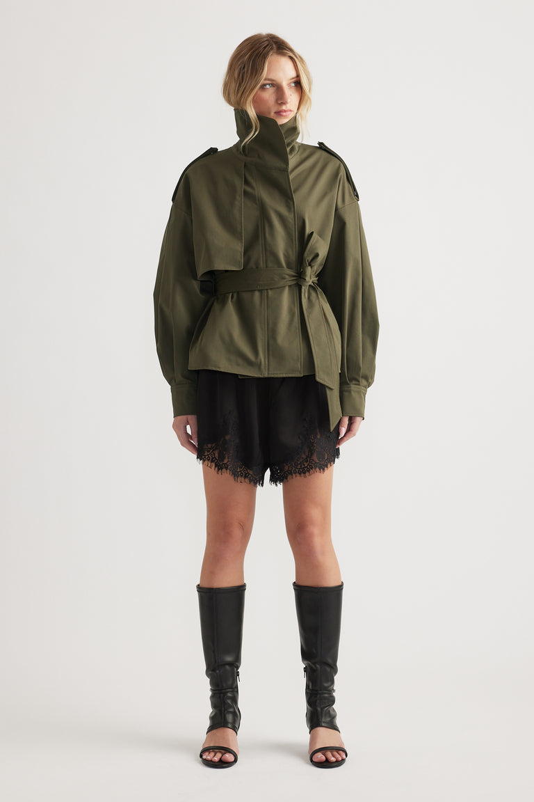 Woman wearing an olive green belted jacket paired with black lace-trimmed shorts and tall black boots