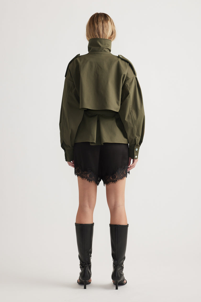 Woman wearing black satin shorts with lace trim paired with an olive green structured jacket and black knee-high boots