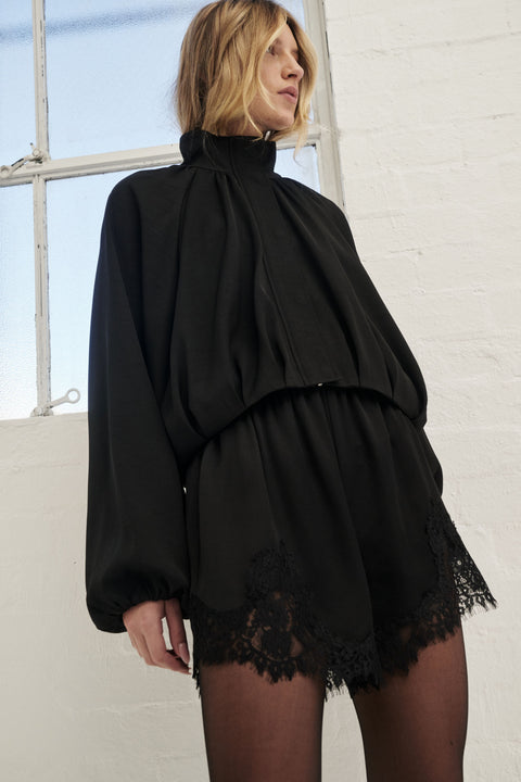 Woman in black satin lace-trim shorts styled with a high-neck blouse and tights
