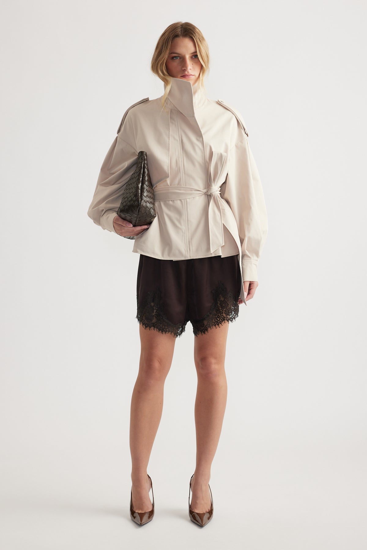 Woman wearing light belted jacket over black lace-trimmed shorts, holding textured clutch and wearing pointed heels