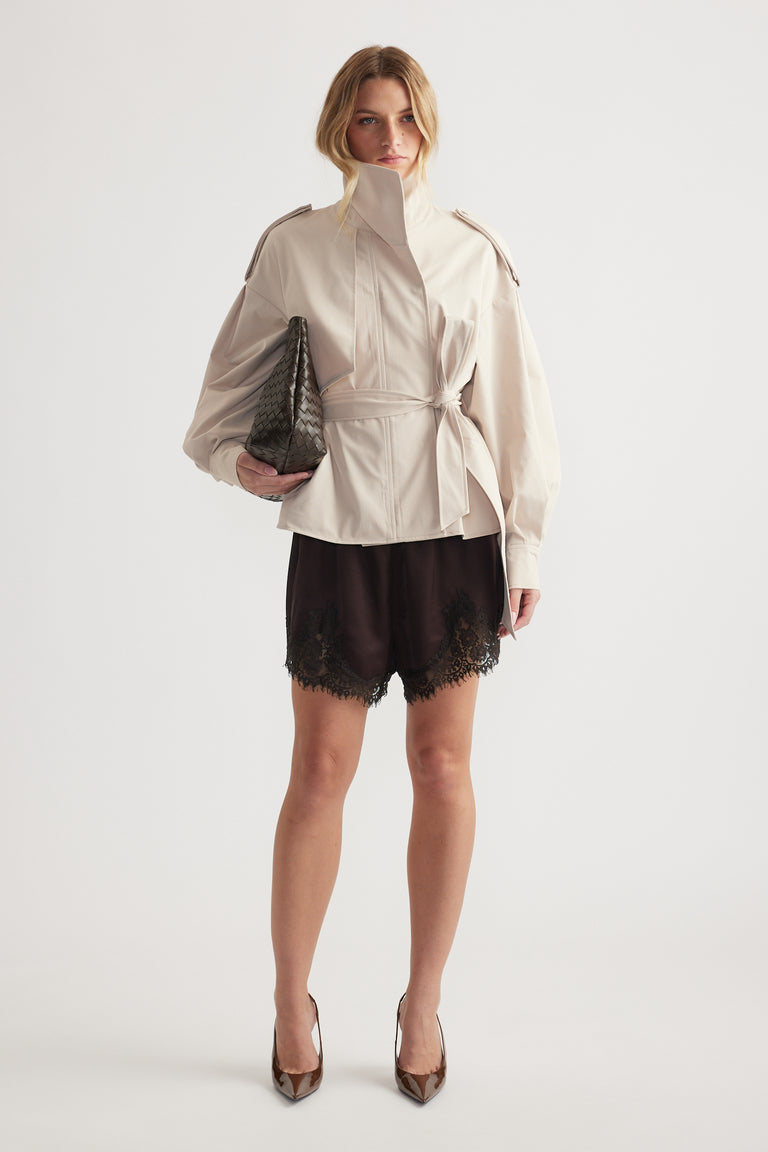 Woman wearing light belted jacket over black lace-trimmed shorts, holding textured clutch and wearing pointed heels
