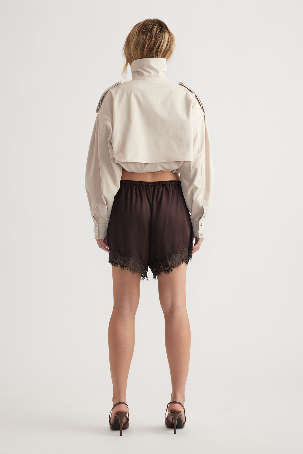 Back view of woman wearing black lace-trimmed satin shorts paired with cropped beige jacket and heels