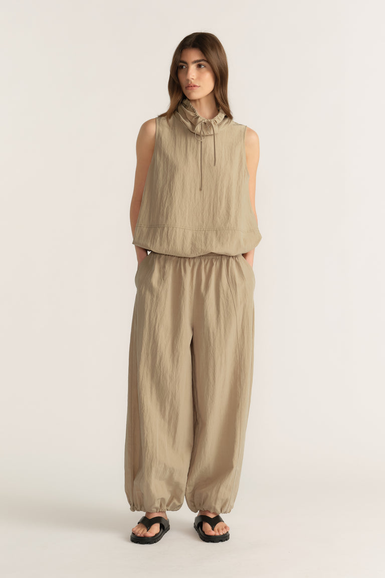 Woman wearing a beige sleeveless funnel neck top with tie detail and matching relaxed pants