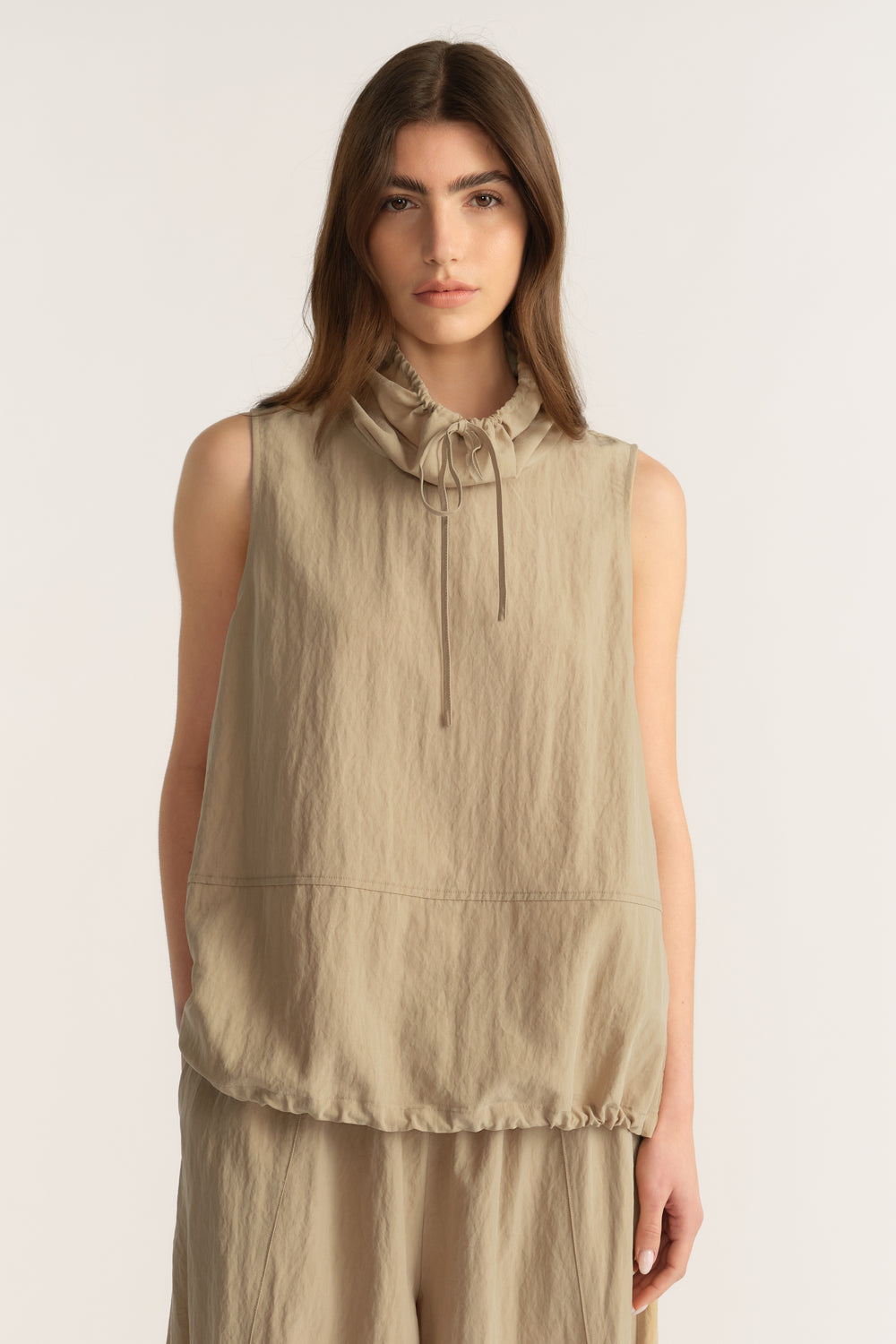 Woman wearing a beige sleeveless funnel neck top with tie details and matching relaxed pants