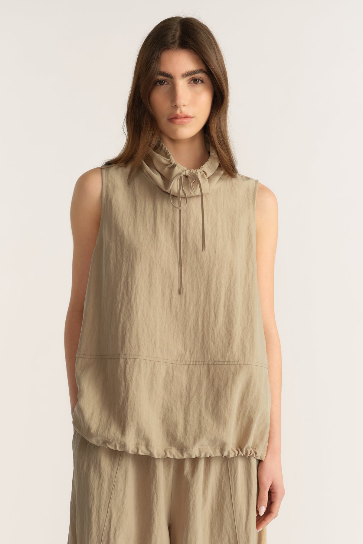Woman wearing a beige sleeveless funnel neck top with tie details and matching relaxed pants