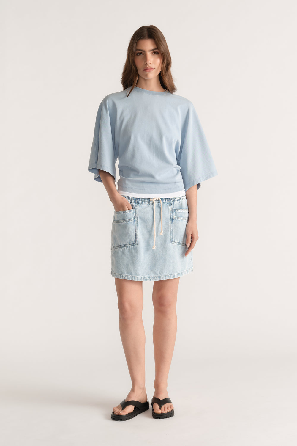 Model wearing a light blue relaxed fit tee with batwing sleeves, a denim mini skirt and black slides