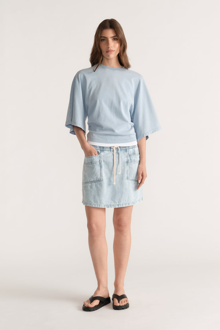 Model wearing a light blue relaxed fit tee with batwing sleeves, a denim mini skirt and black slides