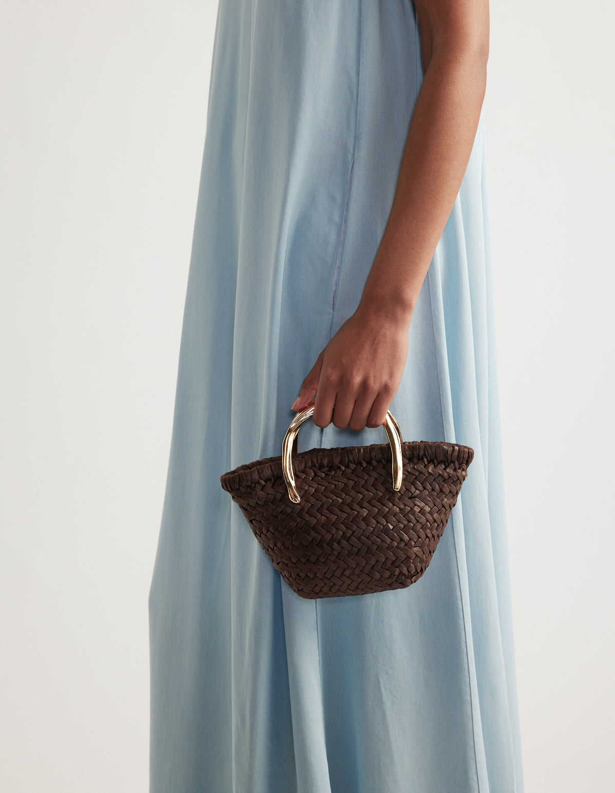 Woman holding a small dark brown paper weave bag with gold metal handles, paired with a pale blue dress