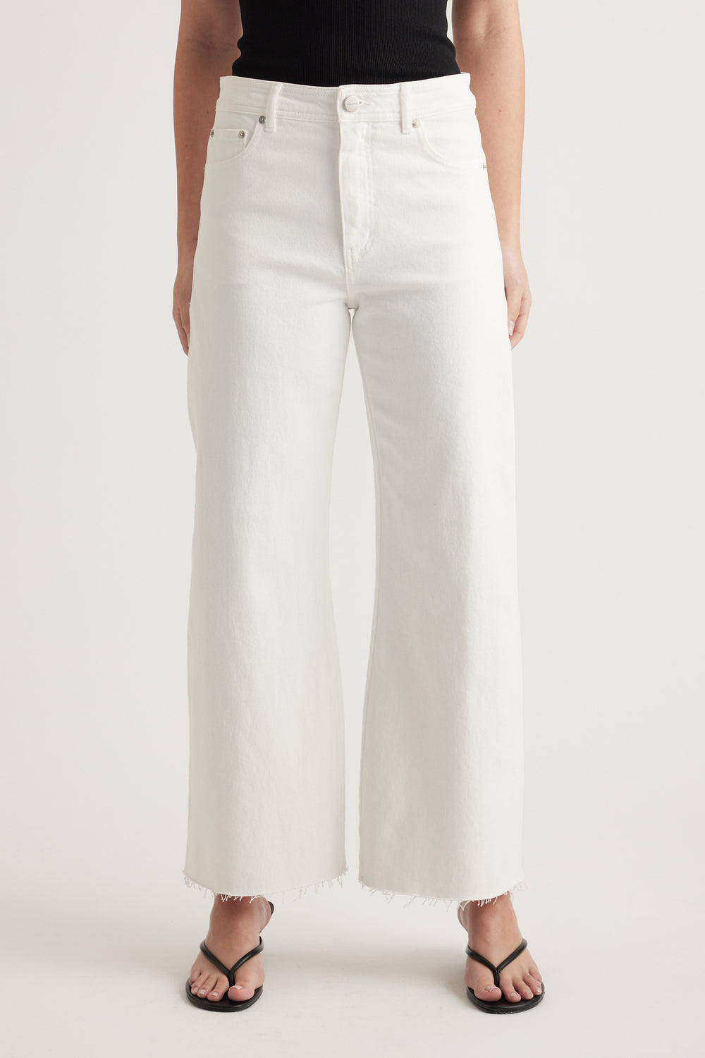 Woman wearing high rise white wide leg jeans with raw hems, a black top and black sandals