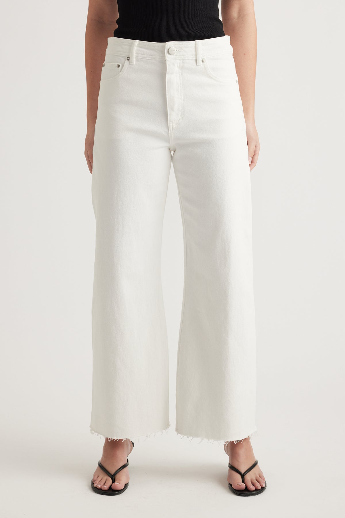 Woman wearing high rise white wide leg jeans with raw hems, a black top and black sandals