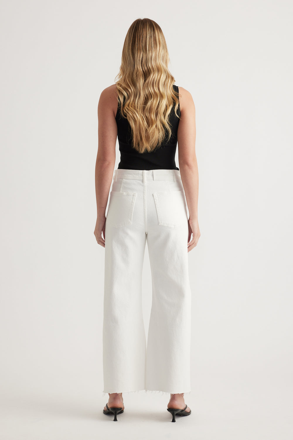 Woman viewed from behind wearing white wide leg jeans with raw hems, a black sleeveless top and heels