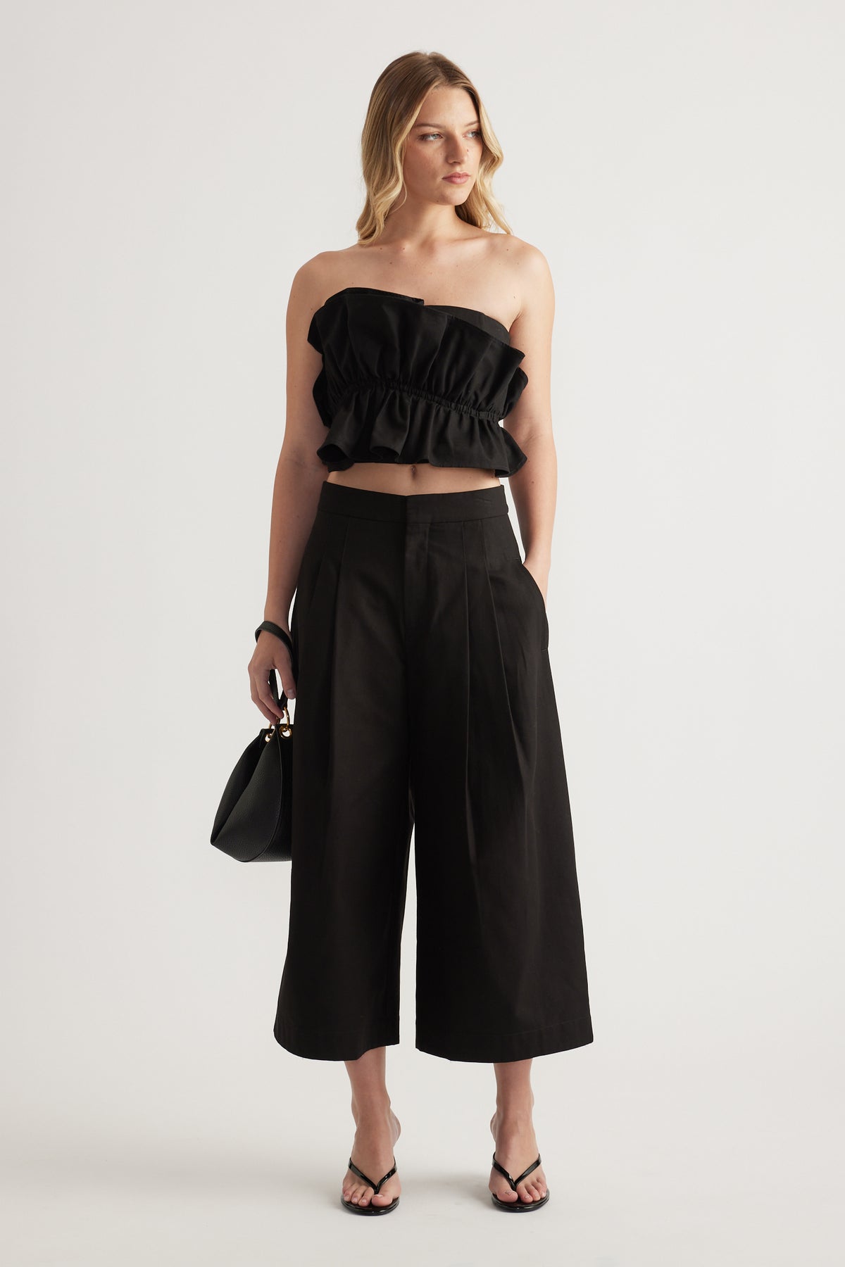 Model wearing a black strapless ruffle top with wide leg pants, handbag and strappy sandals
