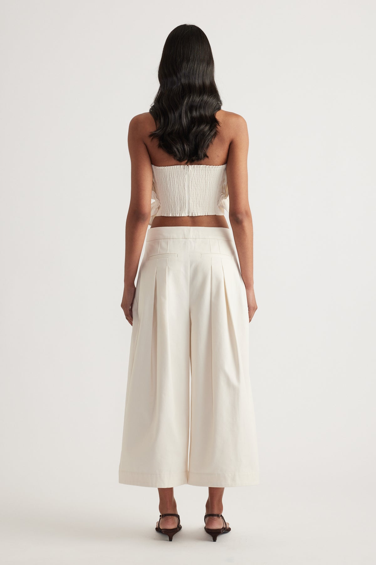 Back view of a woman wearing an ivory strapless cropped top with wide leg pleat pants and black heels