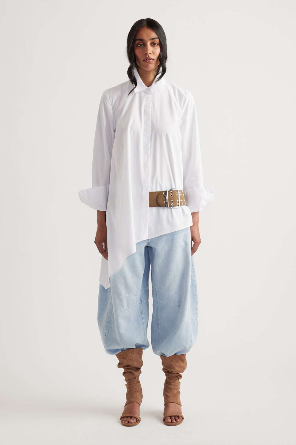 Woman wearing an asymmetric white shirt with a belt detail, paired with light blue baggy pants and tan slouchy boots