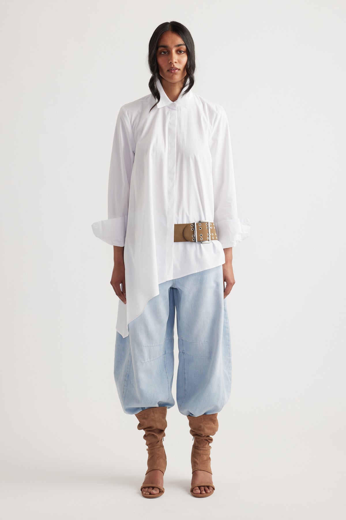Woman wearing an asymmetric white shirt with a belt detail, paired with light blue baggy pants and tan slouchy boots
