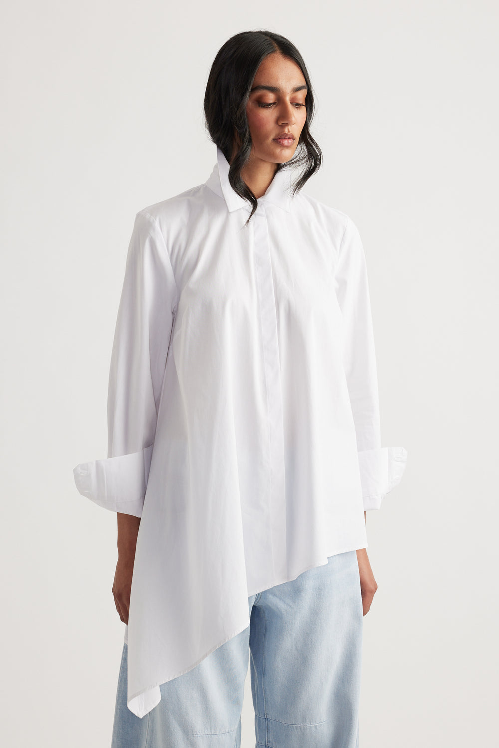 Woman wearing a white asymmetric shirt with a high collar paired with light blue relaxed jeans against a plain background