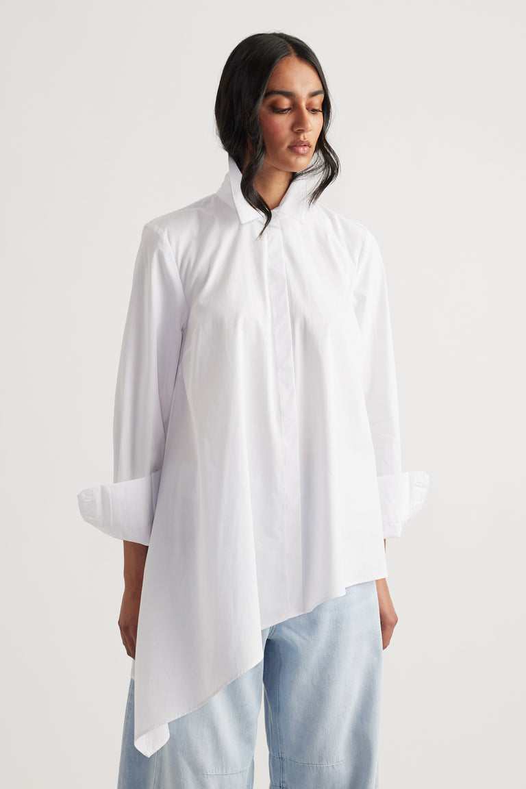 Woman wearing a white asymmetric shirt with a high collar paired with light blue relaxed jeans against a plain background