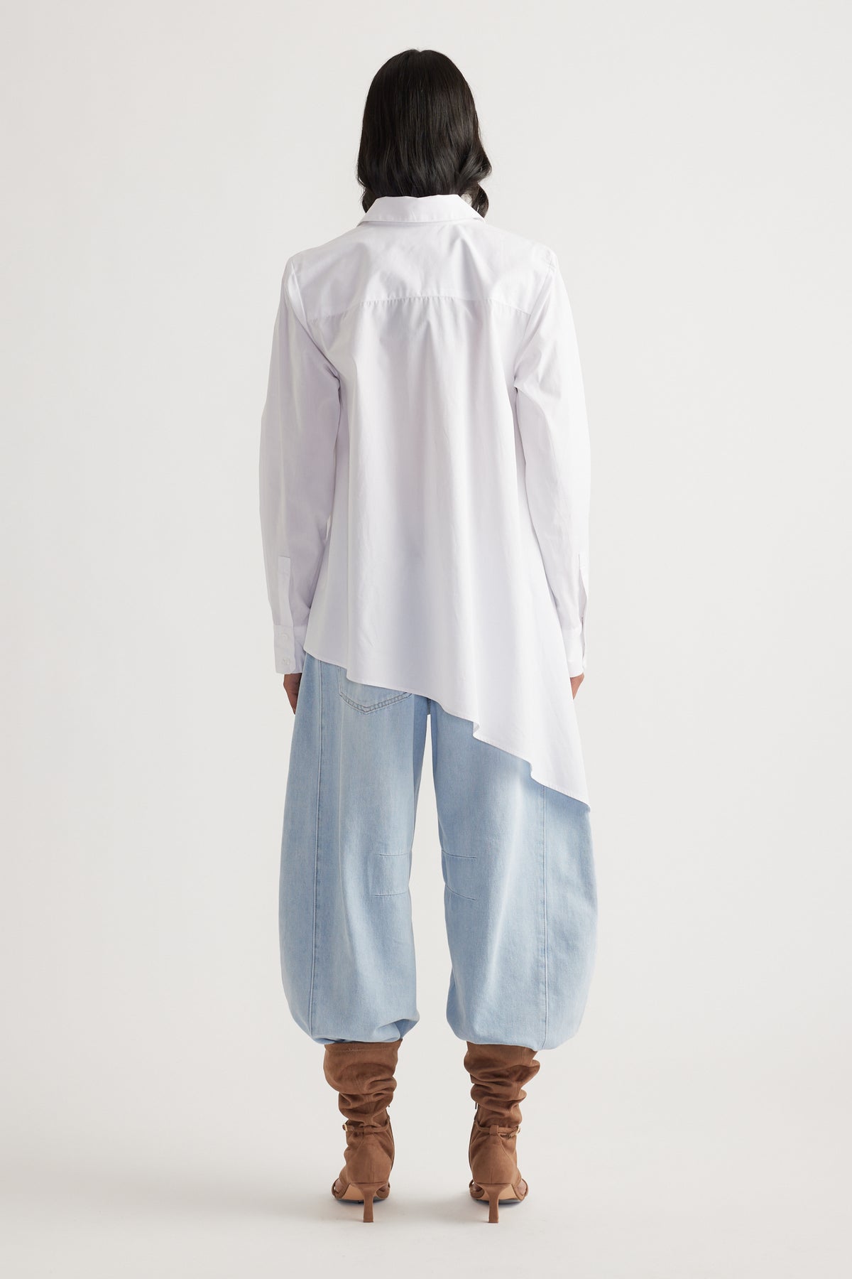 Woman seen from behind wearing a white asymmetric shirt paired with loose light-wash denim pants and slouchy brown boots