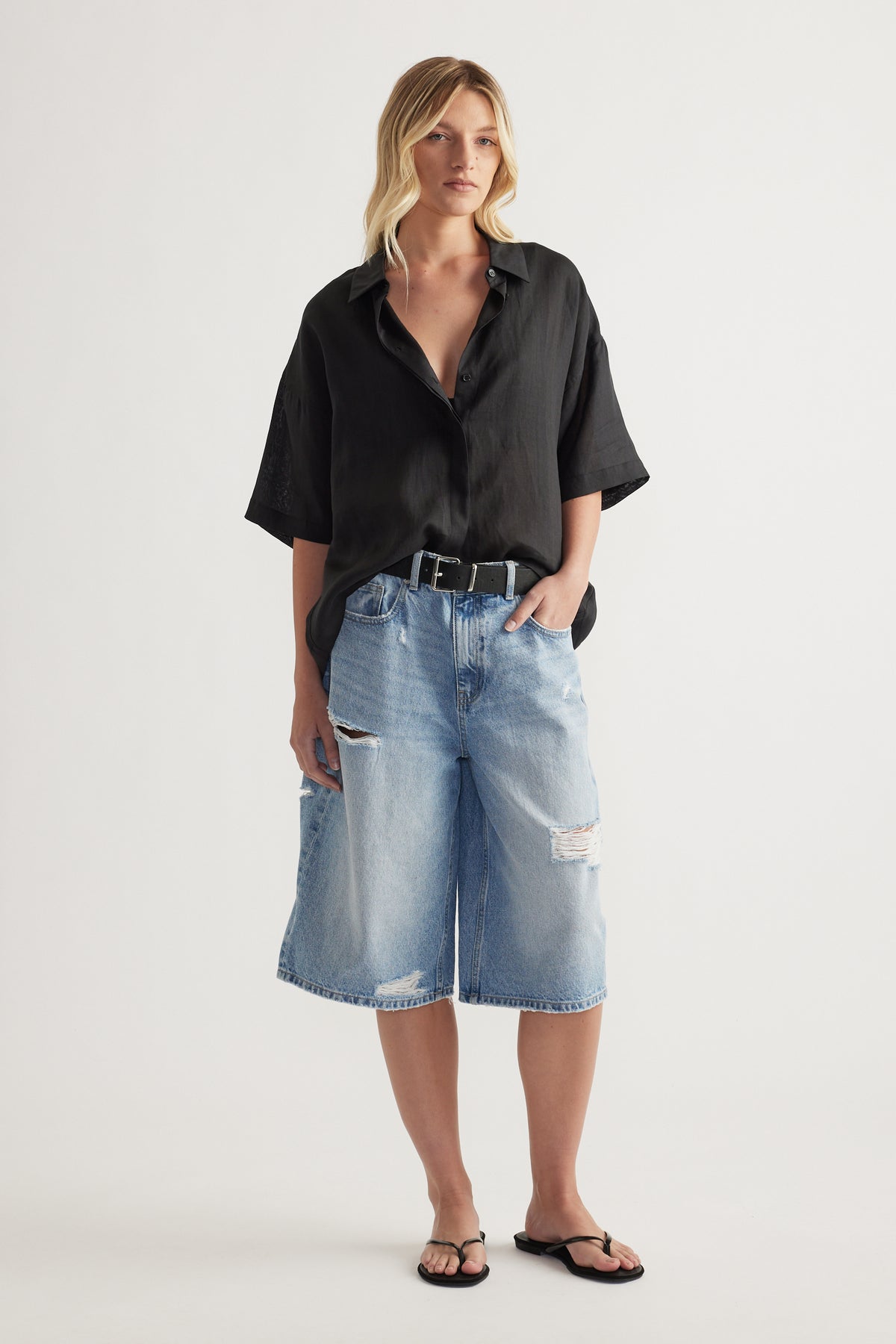 Model wearing a black oversized collared button up shirt with long sleeves and relaxed knee length denim shorts