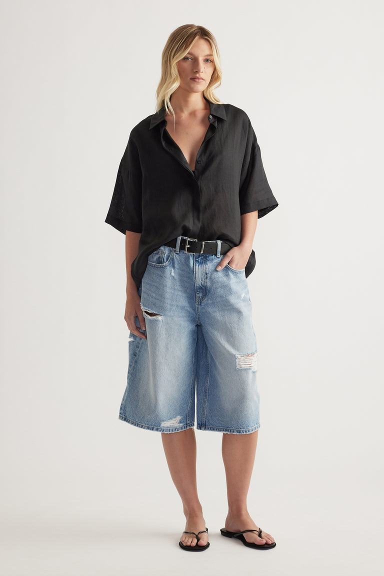 Model wearing a black oversized collared button up shirt with long sleeves and relaxed knee length denim shorts
