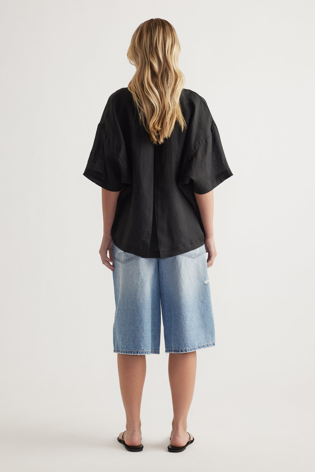 Back view of woman wearing a black oversized button up shirt with elbow length sleeves and light blue denim shorts