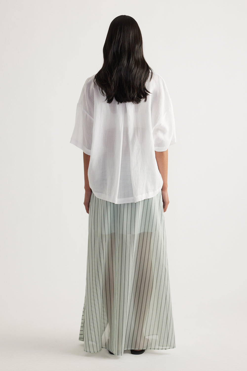 Back view of a woman wearing an oversized white button-up shirt with long sleeves, paired with a pleated maxi skirt