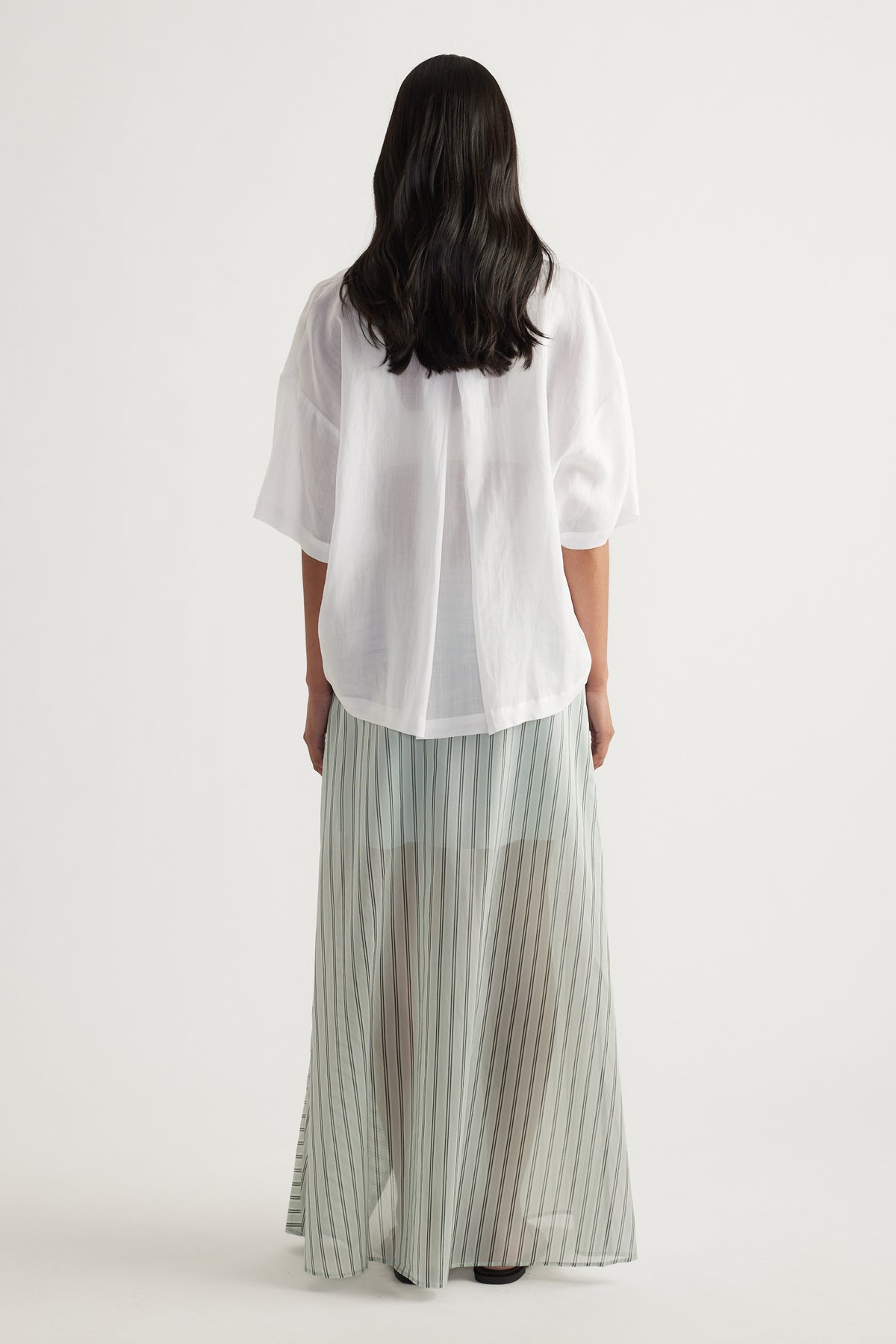 Back view of a woman wearing an oversized white button-up shirt with long sleeves, paired with a pleated maxi skirt