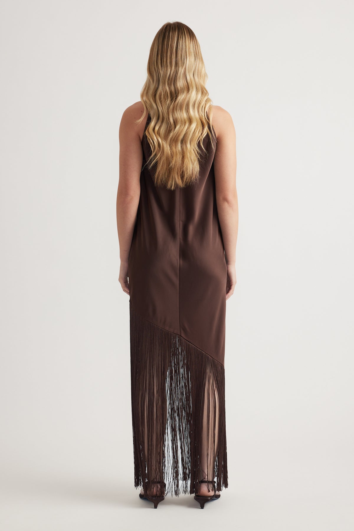 Back view of a woman wearing a brown sleeveless maxi dress with a long fringe hem