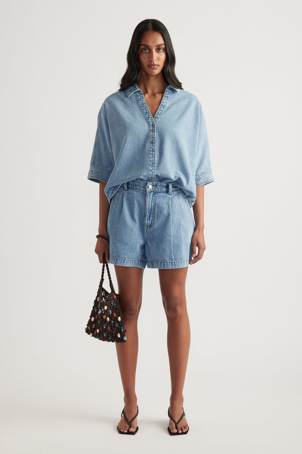 Woman wearing a light denim shirt with high rise denim shorts, black sandals and a beaded handbag