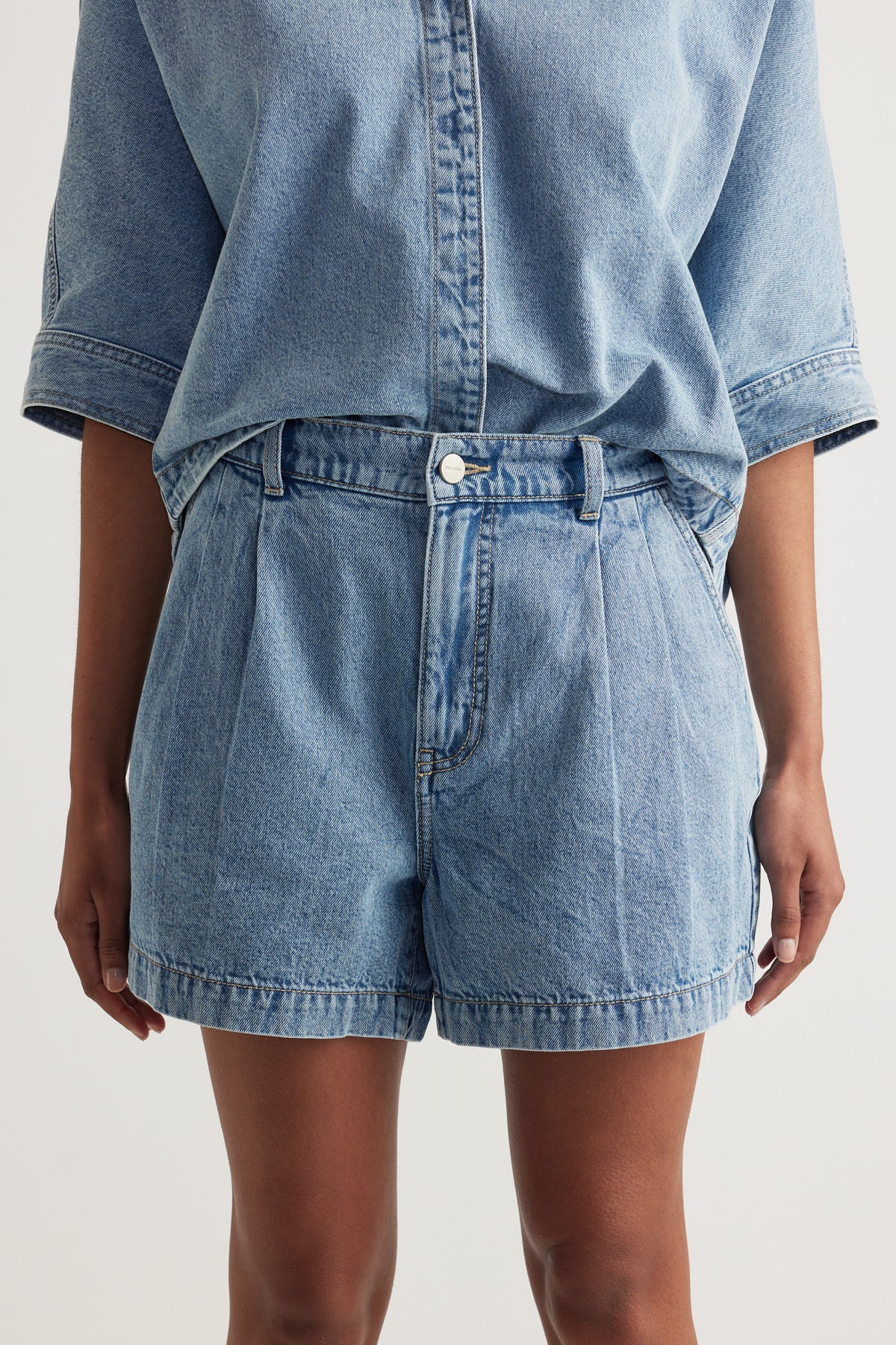 Woman wearing light wash high rise denim shorts with pleat detailing and a matching denim shirt