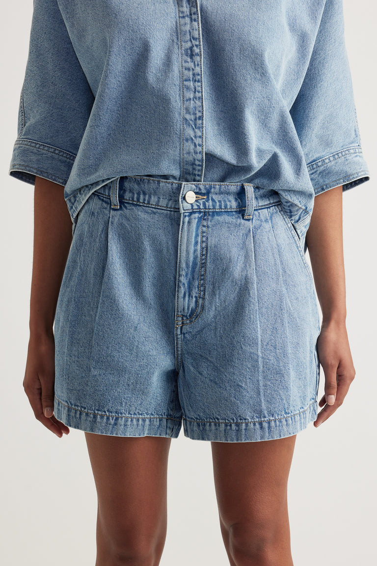 Woman wearing light wash high rise denim shorts with pleat detailing and a matching denim shirt