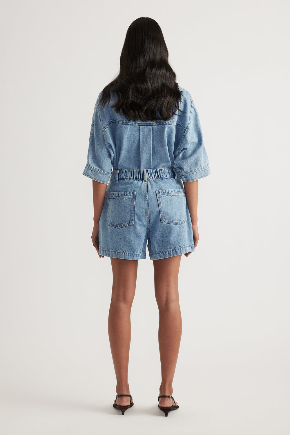 Back view of a woman wearing a light wash denim shirt and high waisted shorts with black heels