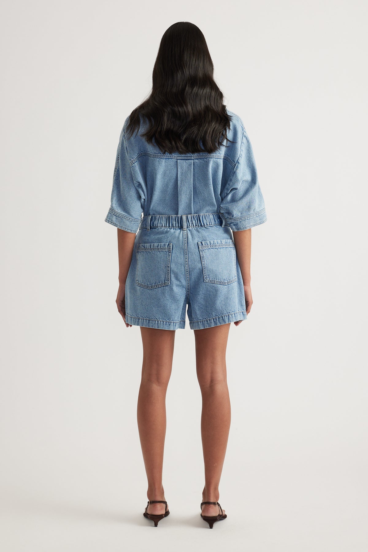 Back view of a woman wearing a light wash denim shirt and high waisted shorts with black heels