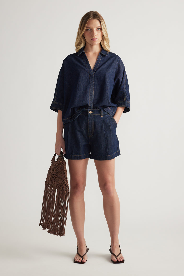 Woman wearing a dark denim shirt with high rise shorts, black sandals and a fringed woven bag