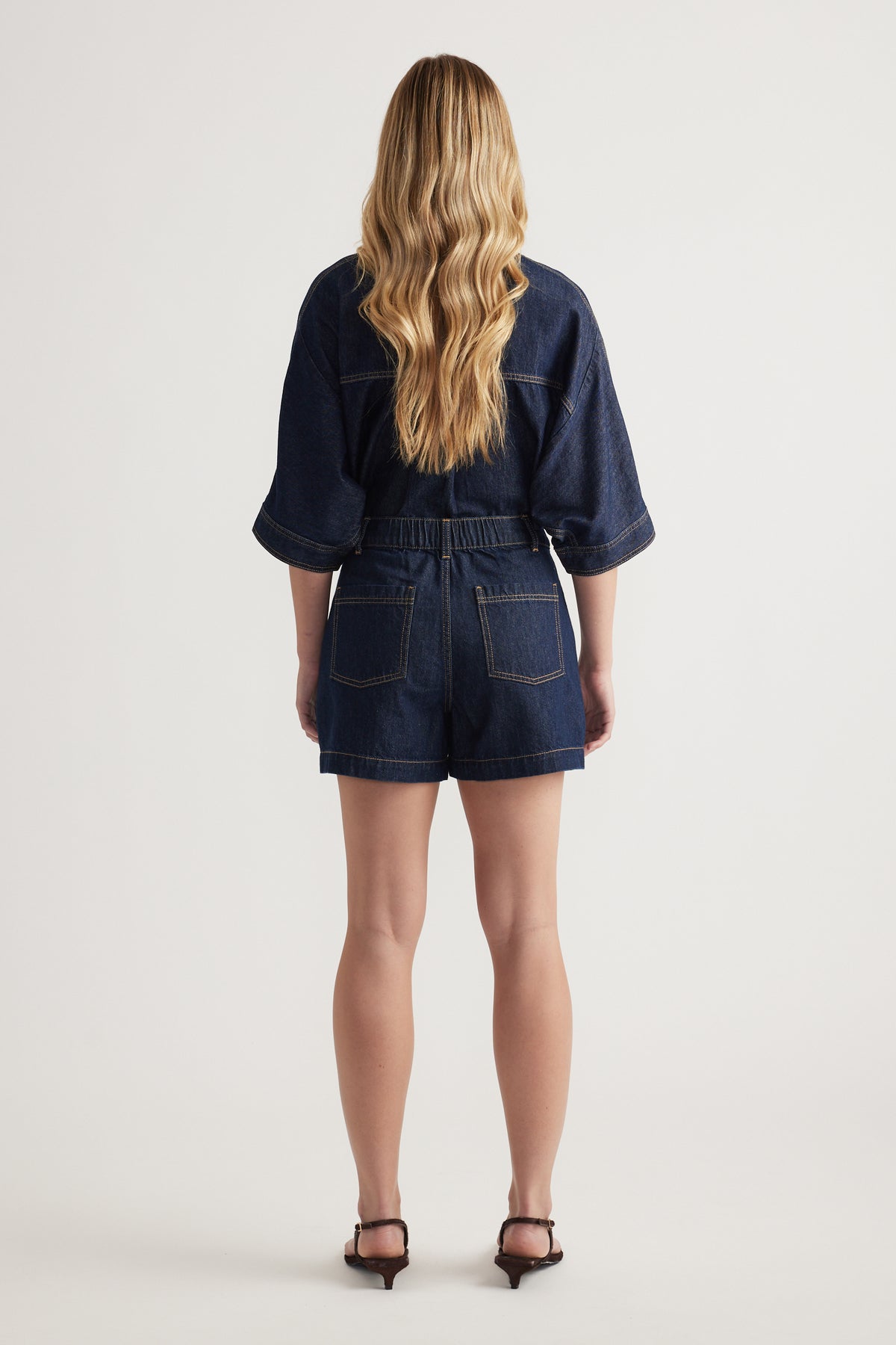Back view of a woman wearing a dark denim playsuit with short sleeves, back pockets and black heels