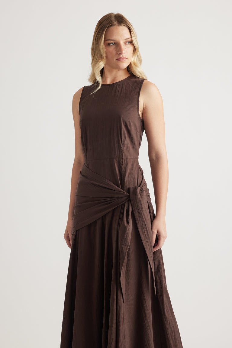 Model wearing a brown sleeveless midi dress with a scarf tie waist and flowing column silhouette