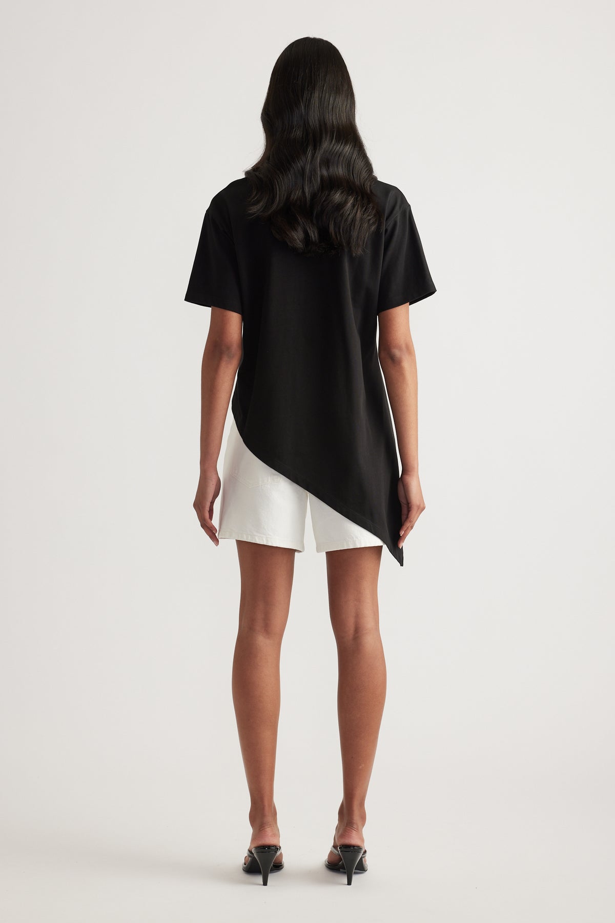 Back view of a woman in a black asymmetric hem tee, white shorts and black heels