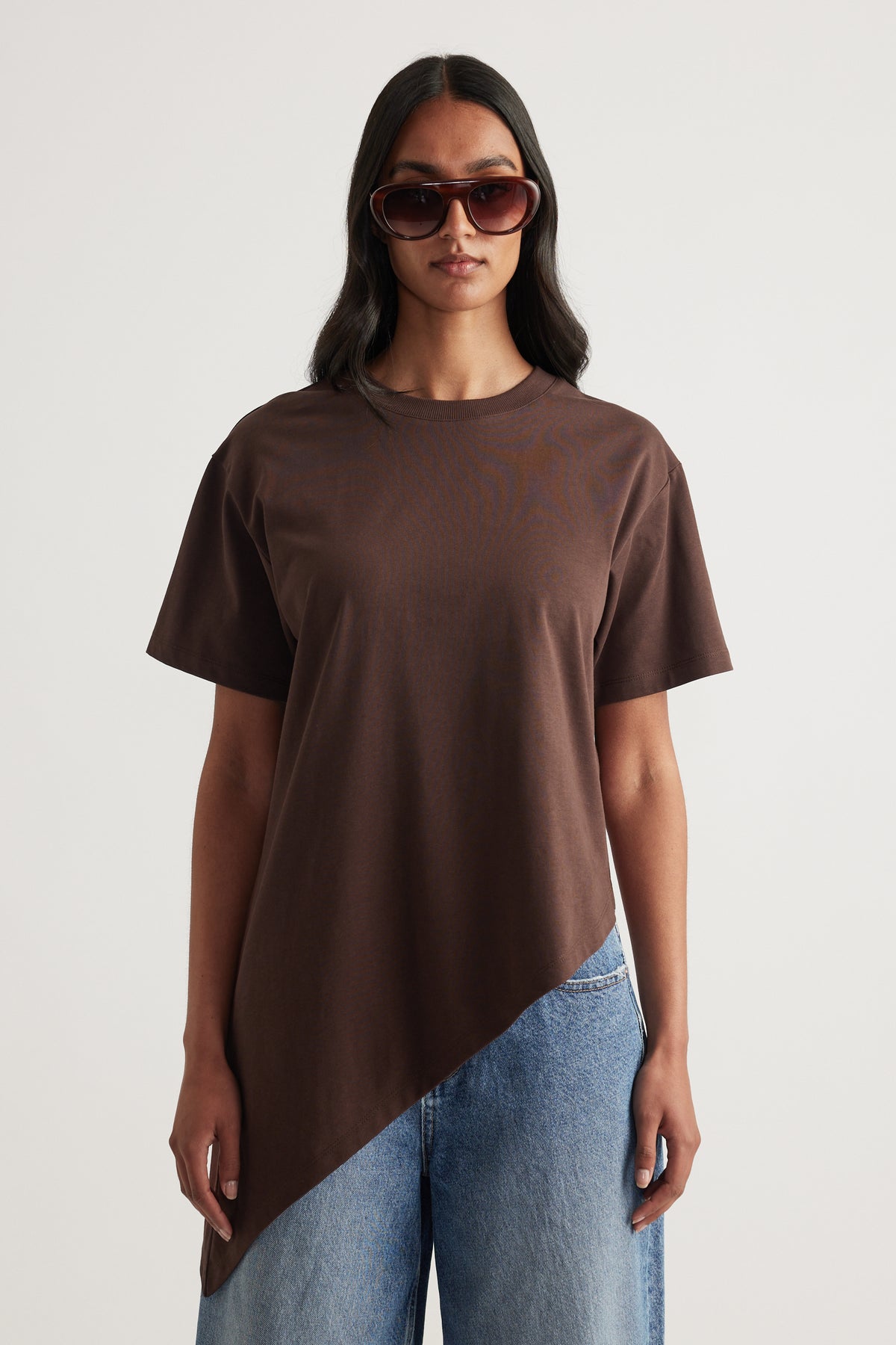 Model in a brown crew neck tee with an asymmetric hem, blue jeans and oversized sunglasses