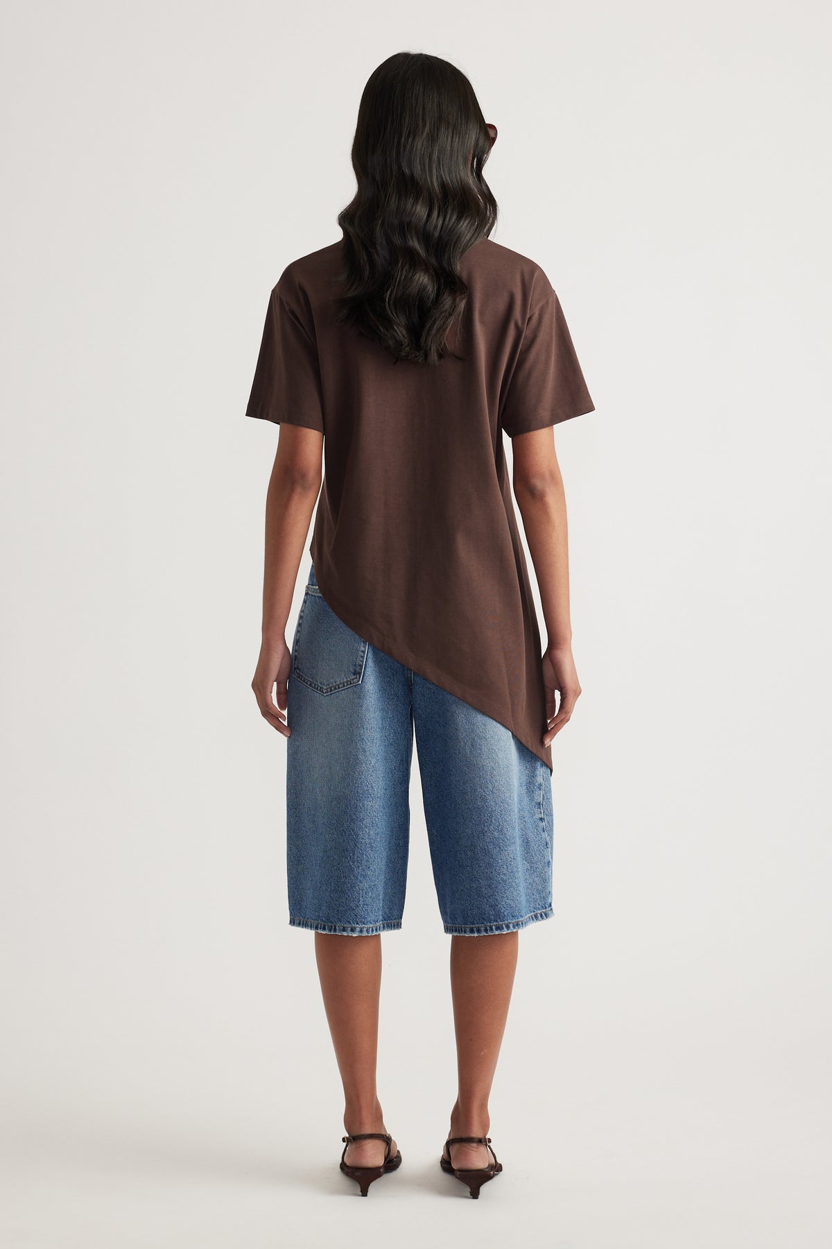 Model wearing a chocolate brown asymmetric hem tee with blue denim shorts and black heeled sandals