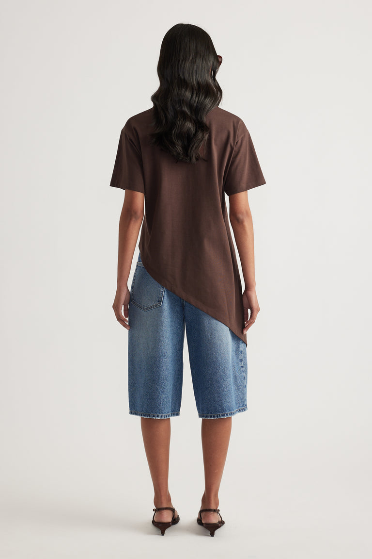 Model wearing a chocolate brown asymmetric hem tee with blue denim shorts and black heeled sandals