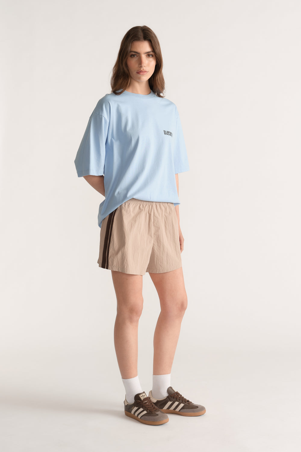Woman wearing beige shorts with dark side stripes, a light blue tee and brown sneakers