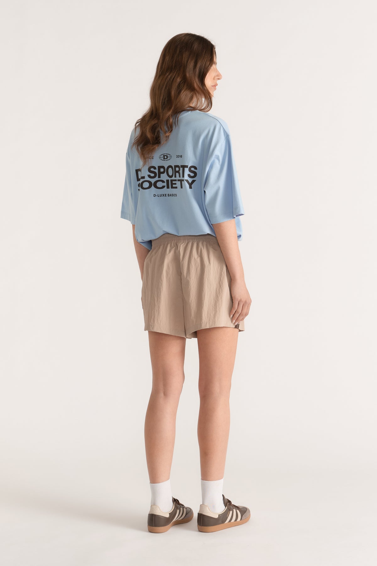 Woman wearing beige elastic waist shorts with a blue oversized tee, white socks and brown sneakers
