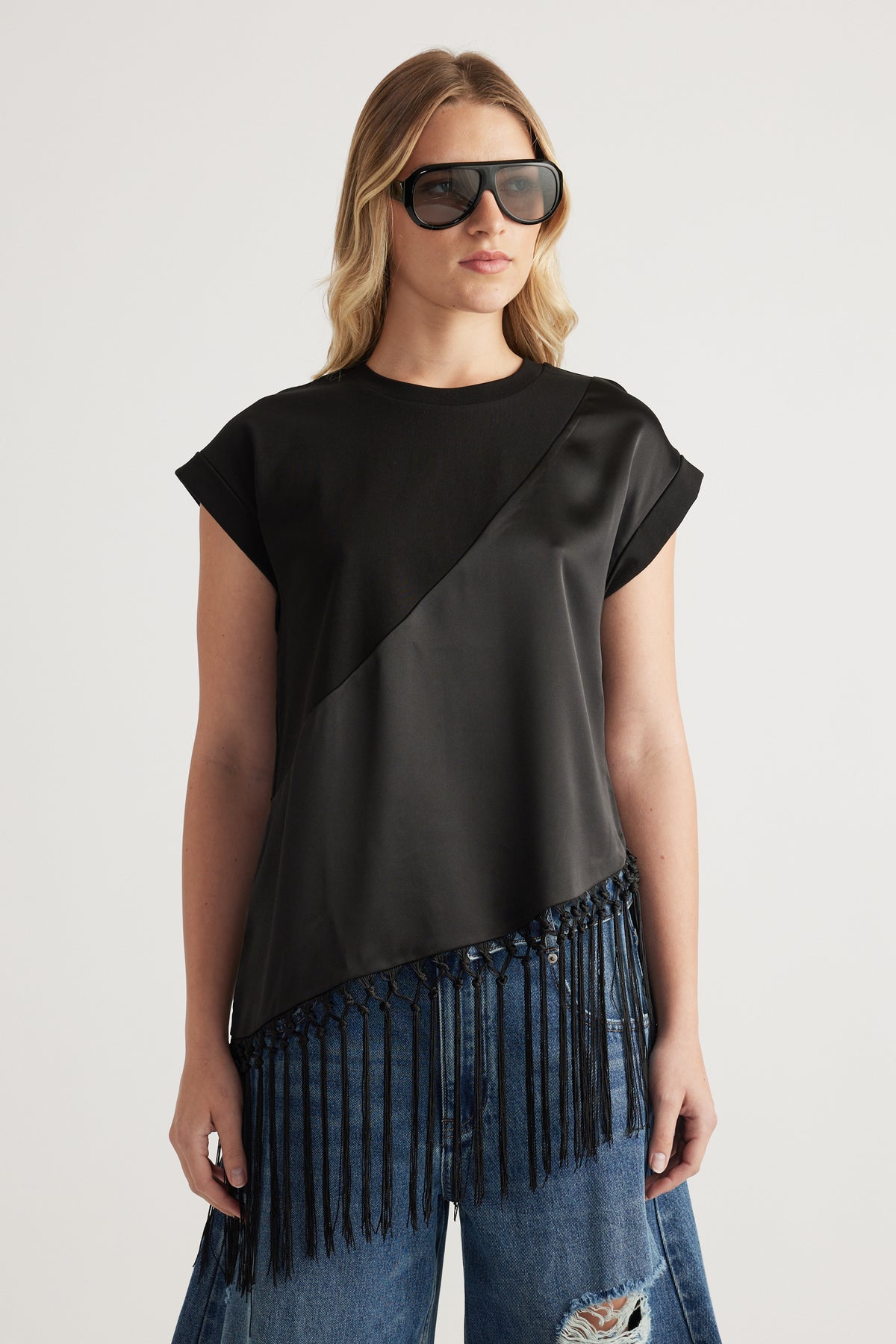 Woman wearing a black tee with satin detail and an asymmetric fringe hem, paired with jeans and sunglasses