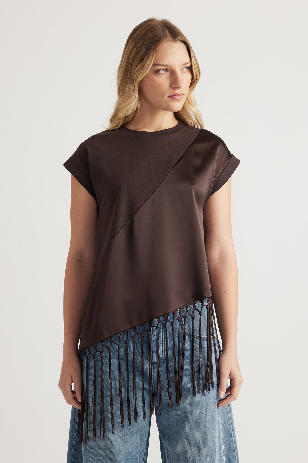 Woman wearing a dark brown asymmetric satin tee with a fringe hem and light wash jeans