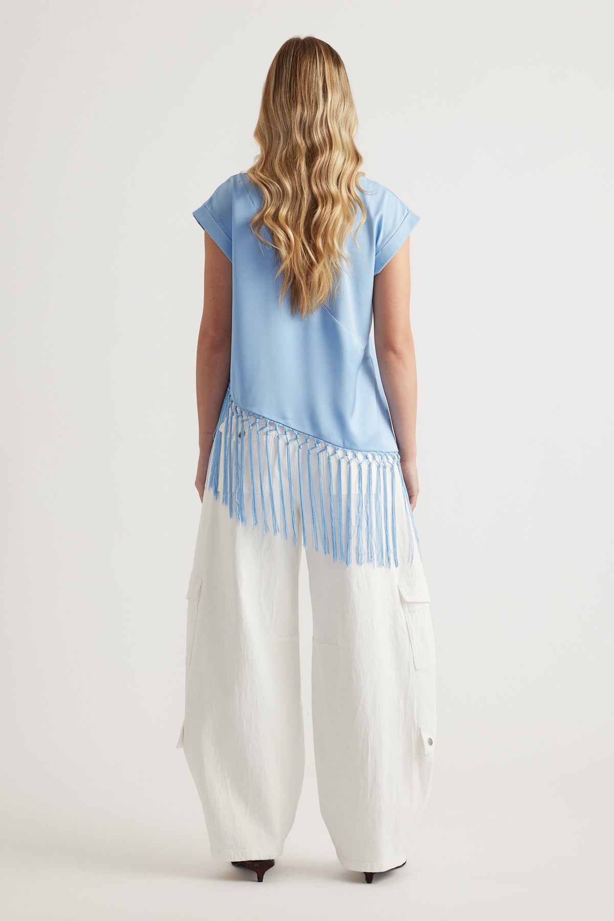 Woman wearing a light blue short sleeve tee with an asymmetric fringe hem and white wide leg pants viewed from behind