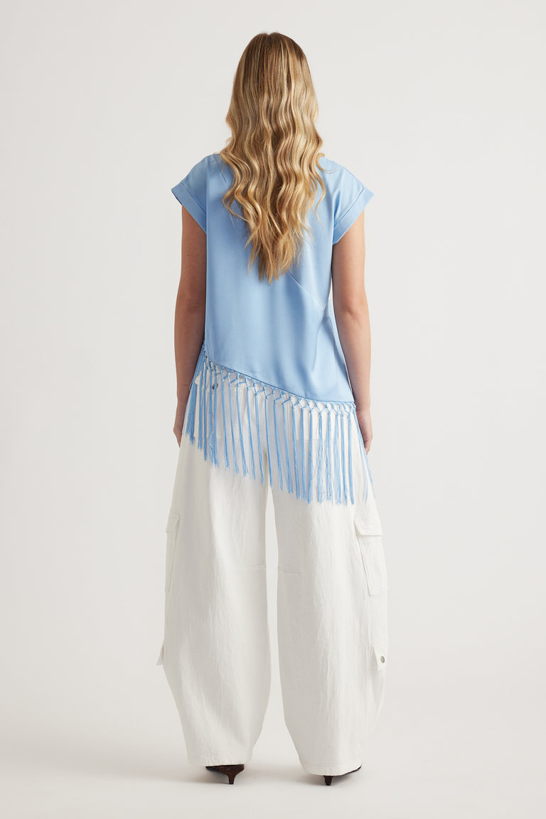 Woman wearing a light blue short sleeve tee with an asymmetric fringe hem and white wide leg pants viewed from behind