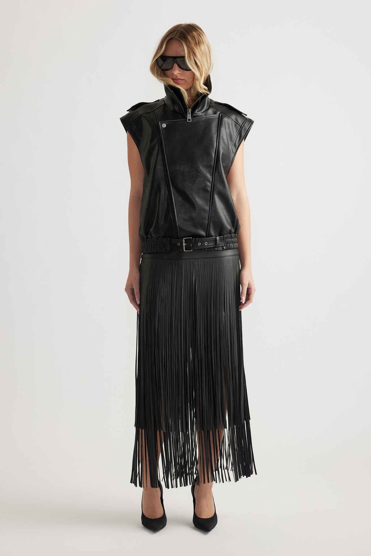 Front view of woman wearing a sleeveless black leather top paired with a high-waisted fringed leather skirt and sunglasses